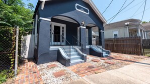 A two-story house with a dark blue exterior, a front porch with steps, and a small front yard with a brick pathway and gravel. - Large Home w/ Back Yard Oasis | Perfect for Groups (New Orleans)