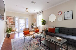 A modern living room with a sofa, coffee table, and chairs. There is a large window with curtains, a plant, and artwork on the walls. - Large Home w/ Back Yard Oasis | Perfect for Groups (New Orleans)