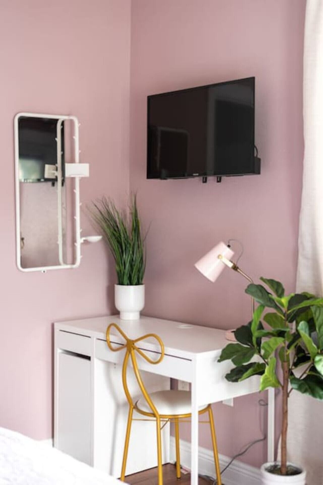 Ein Zimmer mit einer rosafarbenen Wand, einem weißen Schreibtisch mit einem goldfarbenen Stuhl, einem über dem Schreibtisch montierten Fernseher, einem Spiegel an der Wand und einer Topfpflanze auf dem Schreibtisch.