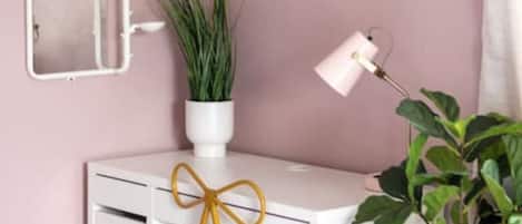 A room with a pink wall, a white desk with a gold chair, a TV mounted above the desk, a mirror on the wall, and a potted plant on the desk.