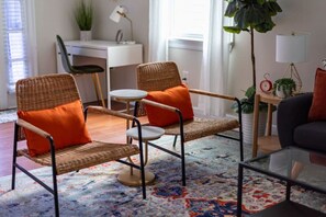 A room with two wicker chairs with orange cushions, a small round table, a large potted plant, and a desk with a chair. - Large Home w/ Back Yard Oasis | Perfect for Groups (New Orleans)