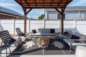 An outdoor patio with a wooden pergola, a gray sofa, black and white chairs, and a woven coffee table.