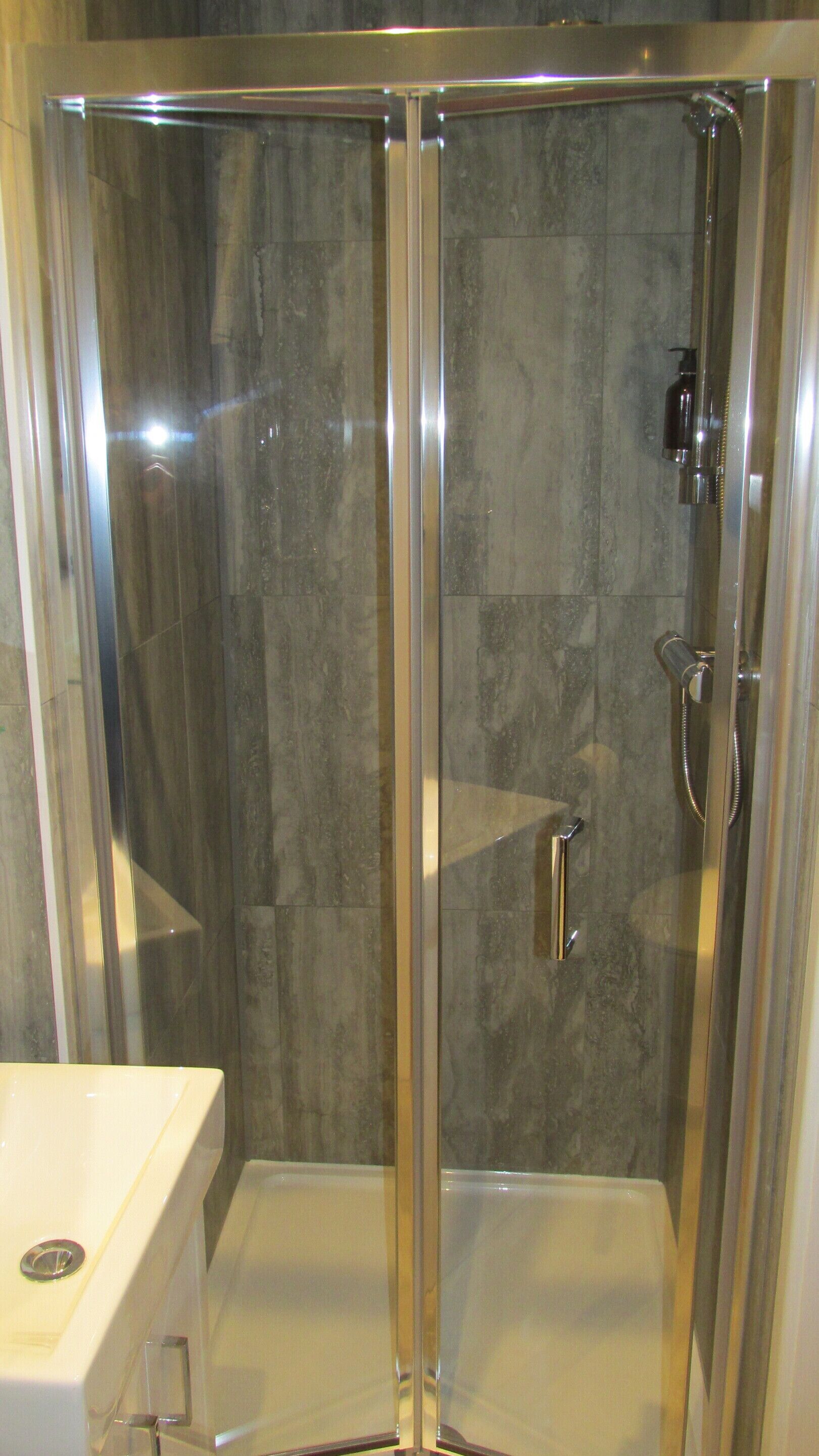 A glass-enclosed shower stall with a handheld showerhead and a bottle of shampoo.