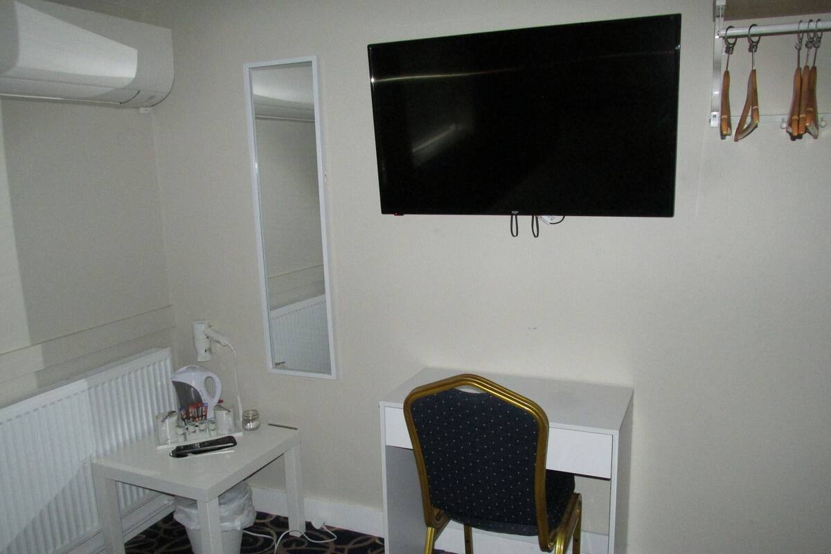 A hotel room with a white desk, a chair, a mirror, a TV, and a radiator.