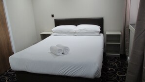 Uma cama bem arrumada com lençóis brancos e duas toalhas enroladas, uma cabeceira escura e uma pequena mesa de cabeceira com abajur.