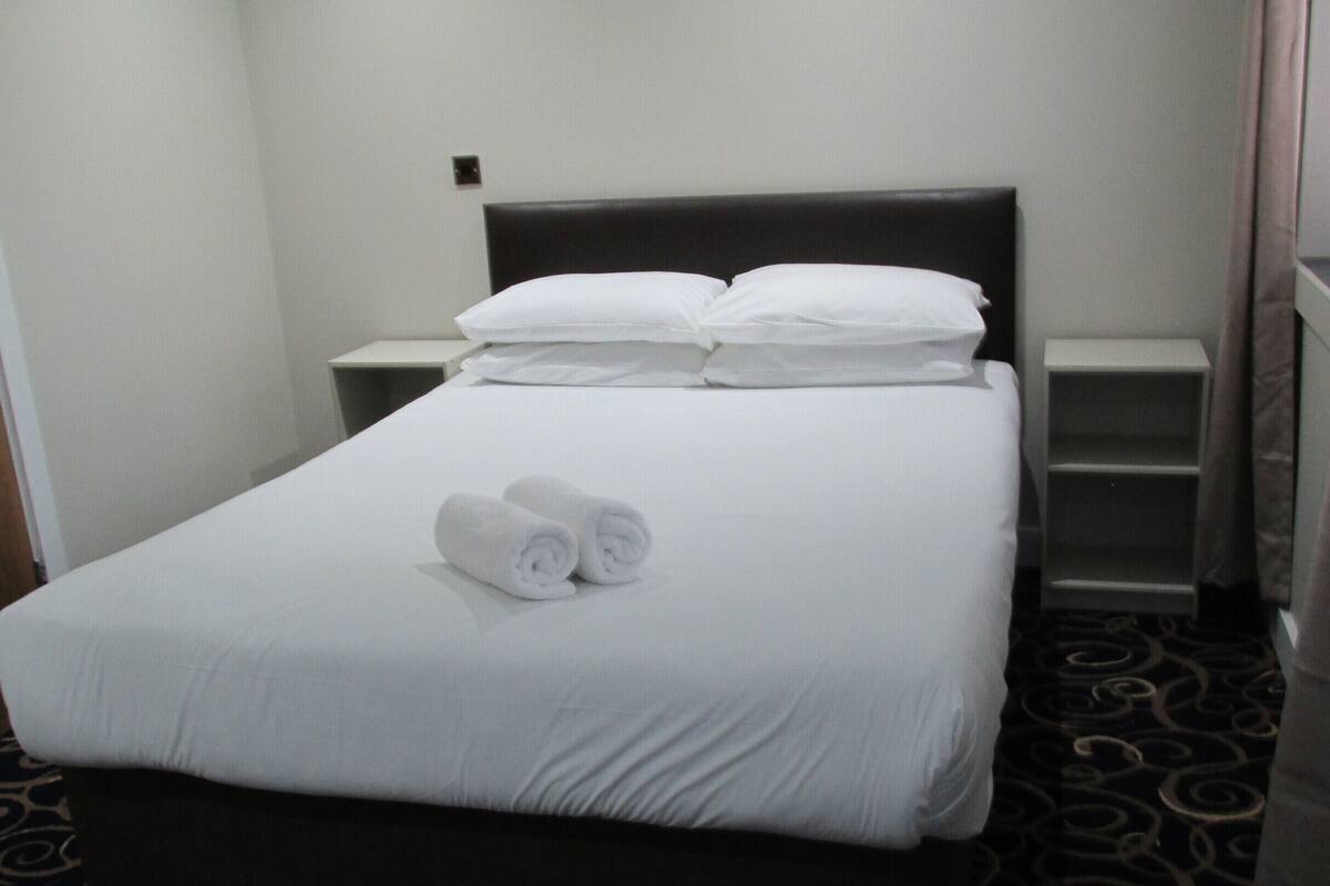 A neatly made bed with white linens and two rolled towels, a dark headboard, and a small bedside table with a lamp.