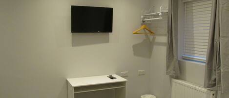 A small, clean bathroom with a white sink, a mirror, and a toilet. There is a black TV mounted on the wall and a window with blinds.