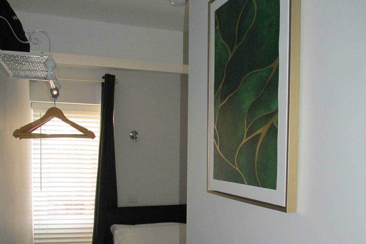 A hotel room with a bed, a hanging light, a ceiling light, a window with blinds, and a framed artwork on the wall.
