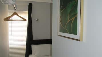 A hotel room with a bed, a hanging light, a ceiling light, a window with blinds, and a framed artwork on the wall.