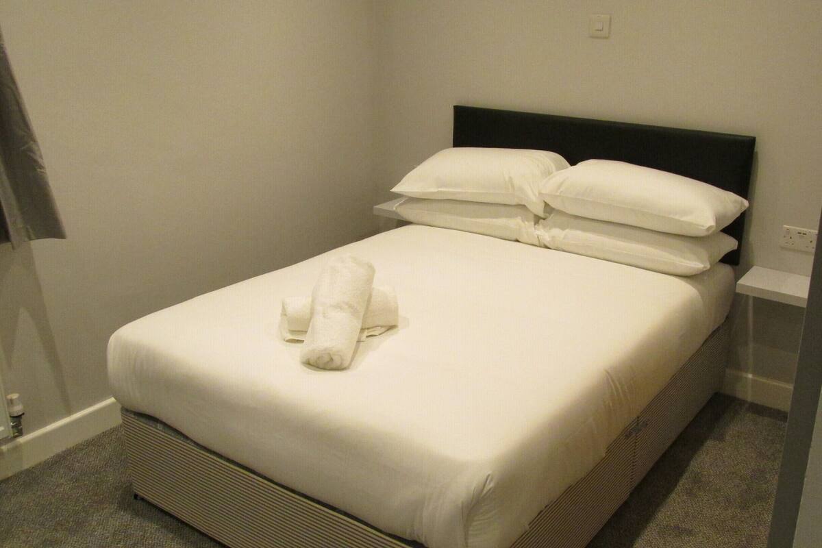 A neatly made bed with white linens and a rolled towel, a black headboard, and a small bedside table with a power outlet.