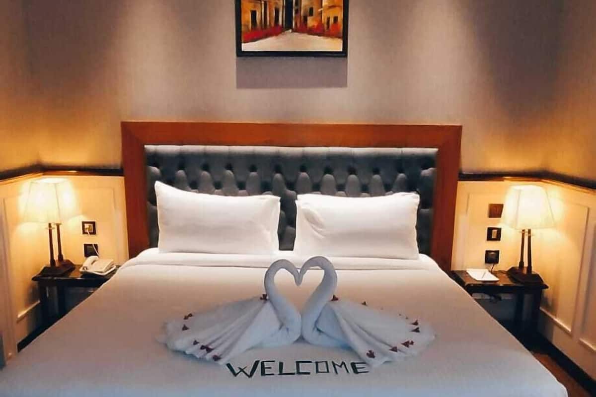 A hotel room with a large bed, two bedside tables with lamps, a framed painting on the wall, and a 'WELCOME' sign on the bed.