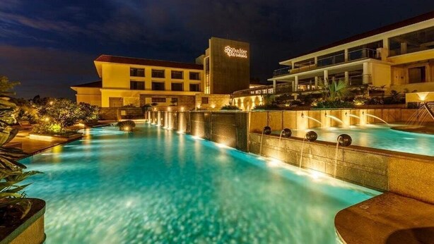 A hotel with a large outdoor swimming pool, a well-lit building, and a landscaped area with plants.