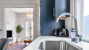 A modern kitchen with a large sink, a coffee maker, and a toaster. There is a dining area with a table and chairs, and a plant in the corner.