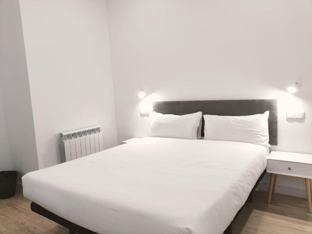 Una cama bien tendida, con ropa blanca y un cabecero de madera. Hay una mesita de noche blanca con un cajón y una lámpara pequeña encima.