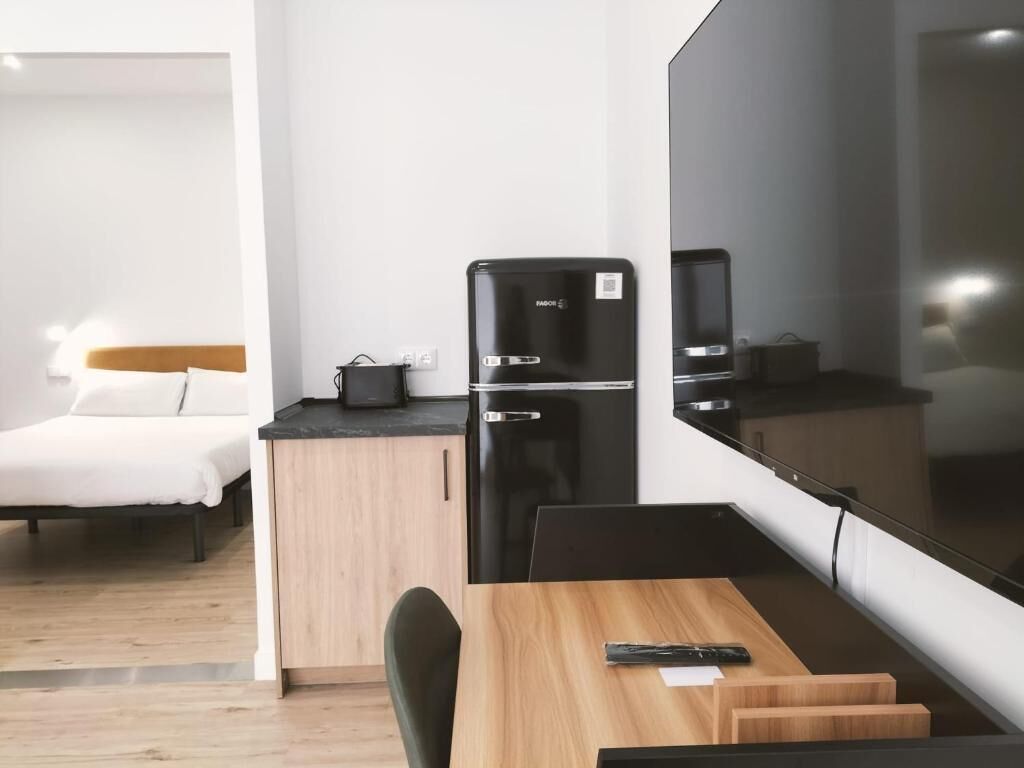 A compact hotel room with a bed, a small kitchenette, a dining area, and a large mirror.