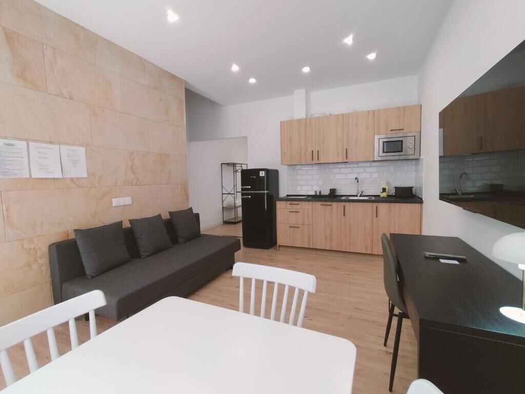 A modern kitchen with wooden cabinets, a black refrigerator, and a microwave. There is a dining area with a white table and chairs.