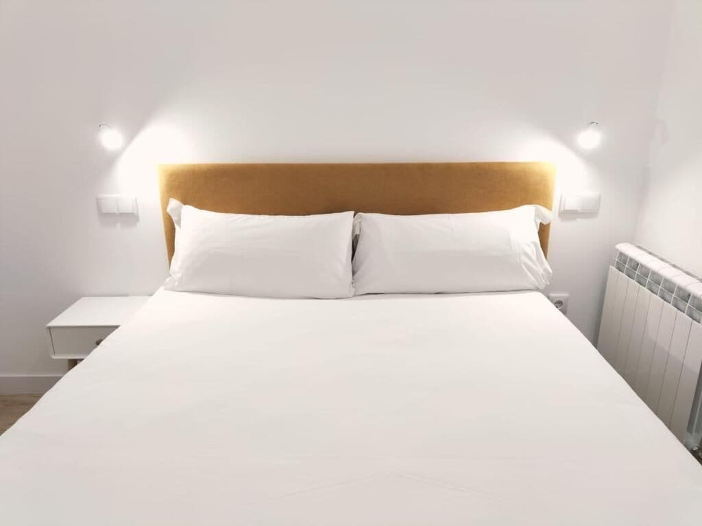 Een net opgemaakt bed met een houten hoofdbord, witte beddengoed en twee wandlampen aan weerszijden.