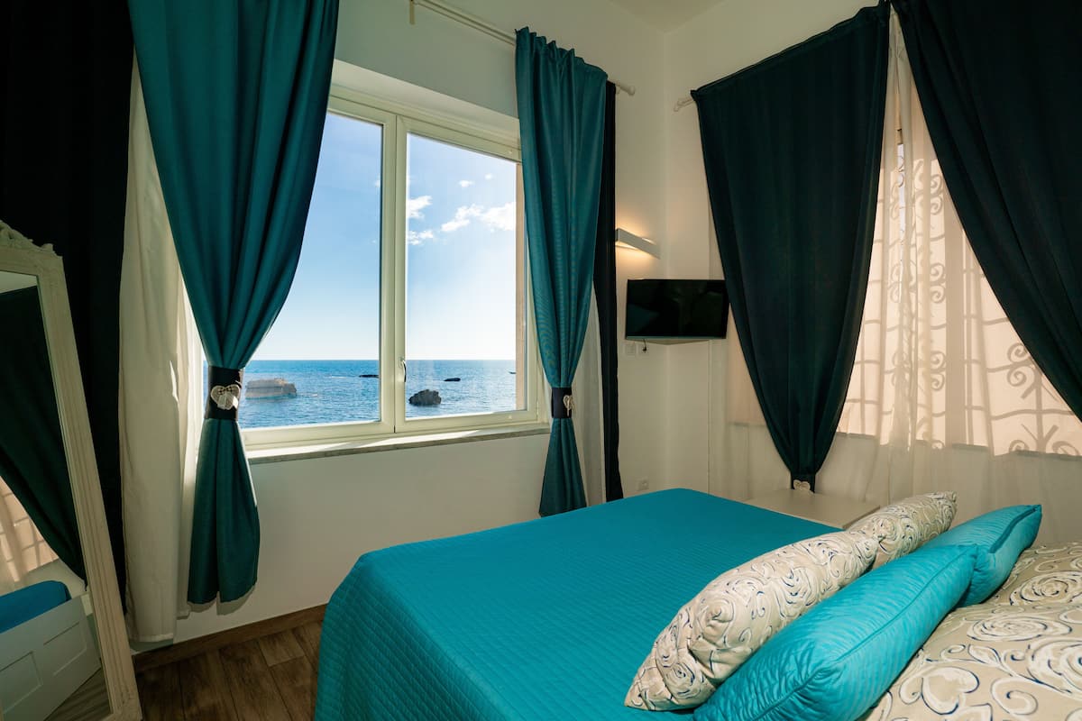 A bedroom with a bed, curtains, a TV, and a view of the sea.