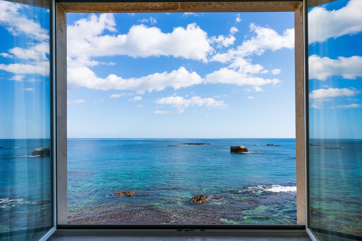 A view through a window of a coastal scene with clear blue water, scattered rocks, and a partly cloudy sky.