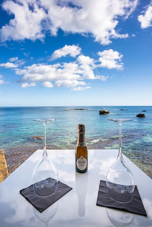 Una mesa con dos copas de vino y una botella de Maschio Pinot Grigio, sobre un mantel blanco, con el mar azul y el cielo despejado de fondo.
