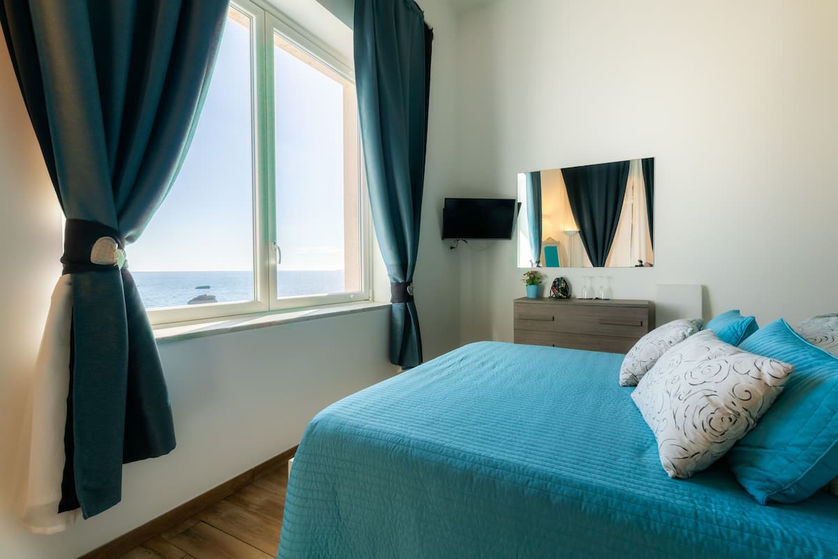 A bedroom with a bed, curtains, a TV, a dresser, and a view of the sea.