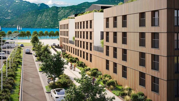 A modern multi-story building with a flat roof and large windows, located near a body of water and mountains.