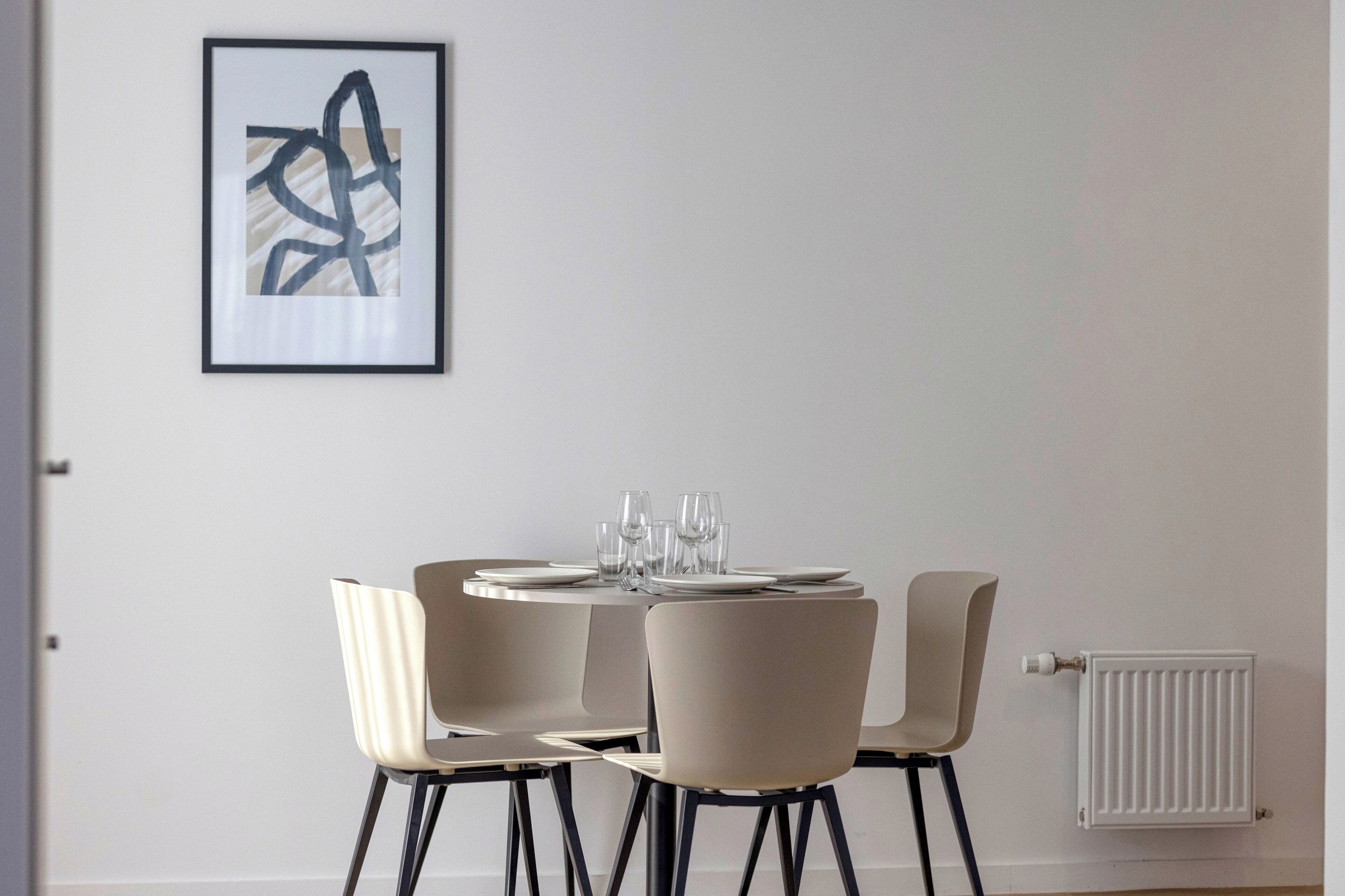 A dining area with a round table set for four, four chairs, and a framed abstract painting on the wall.