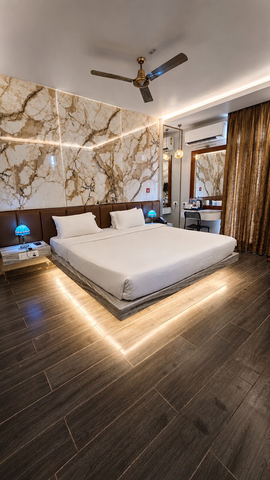 A modern hotel room with a large bed, a ceiling fan, marble-patterned walls, and a wooden floor.