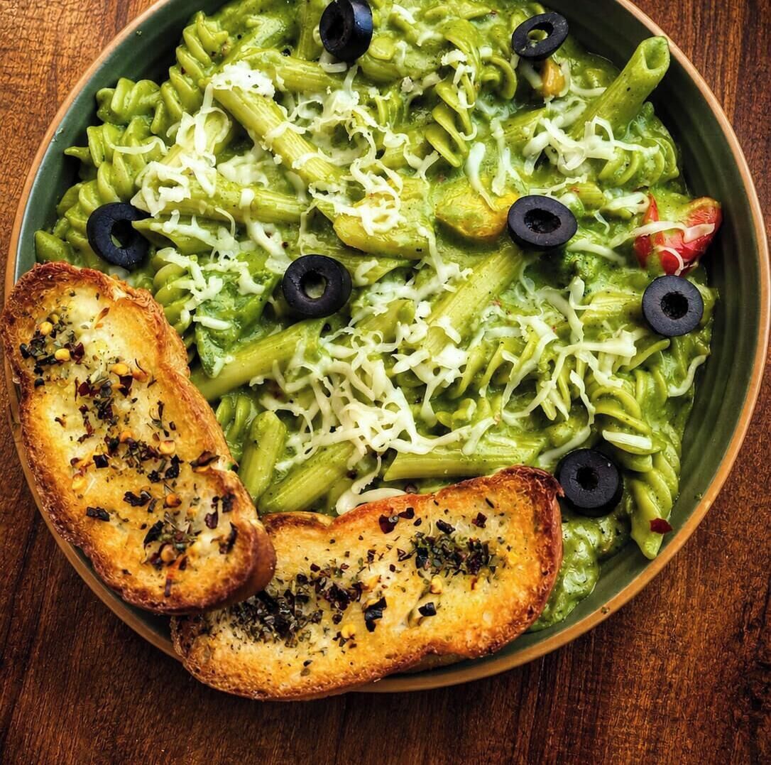 Un plat de pâtes nappées d’une sauce verte, garni d’olives noires et de fromage râpé.