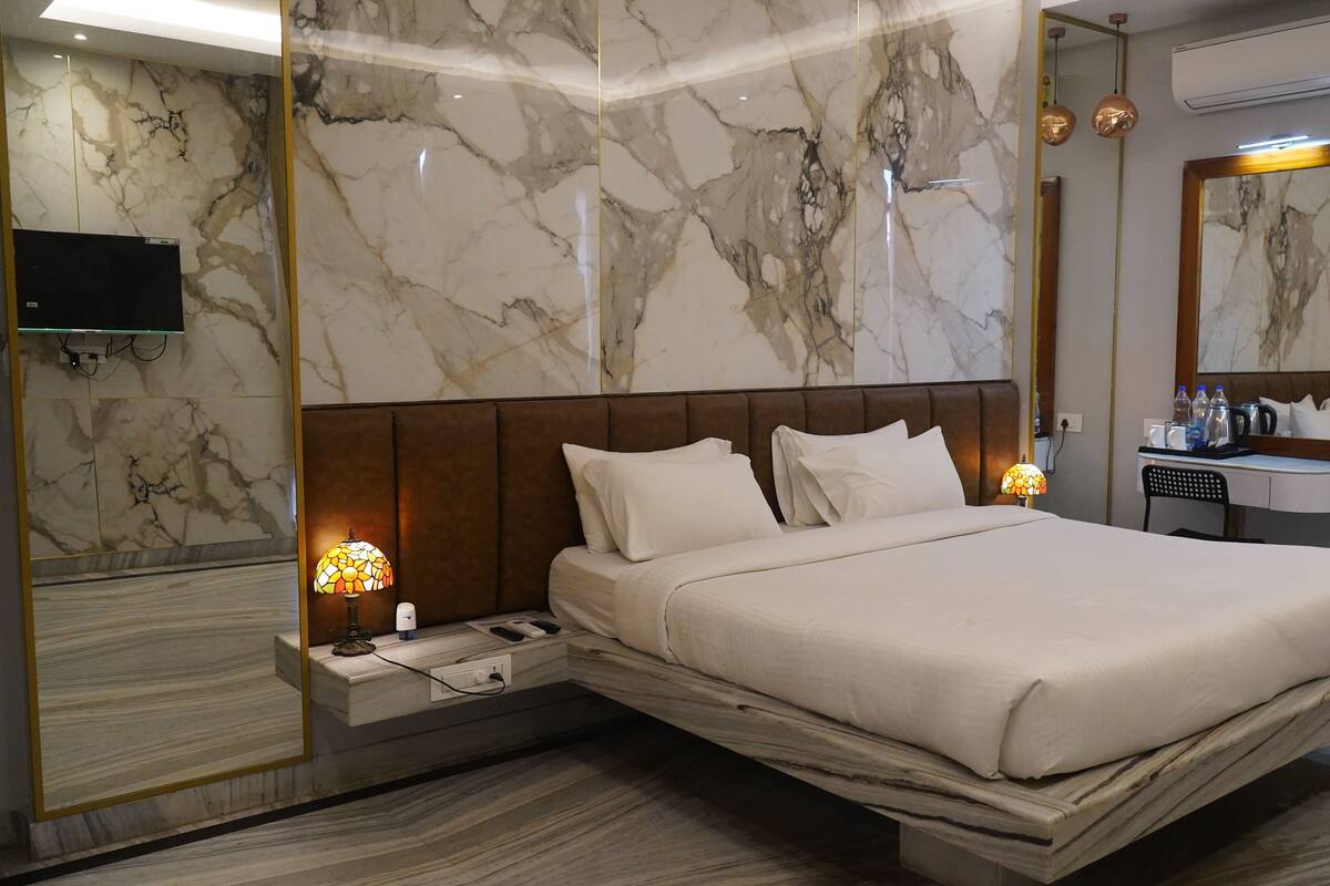 Una habitación de hotel moderna con pared de mármol, una cama grande, una mesita de noche con lámpara, un espejo y un pequeño escritorio con artículos de aseo.