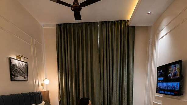 Habitación de hotel con cama, ventilador de techo, televisor y un ventanal con cortinas.