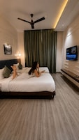 Habitación de hotel con cama, ventilador de techo, televisor y un ventanal con cortinas.