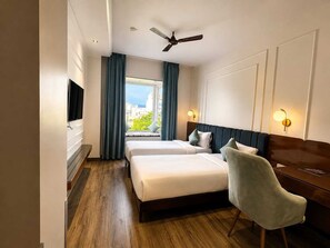 Ein Hotelzimmer mit zwei Betten, einem Schreibtisch, einem Sessel, einem Fernseher, einem Deckenventilator und Blick auf die Stadt.