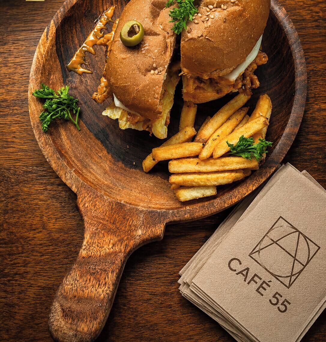 Ein Holzbrett mit einem Sandwich und Pommes, sowie ein Servietttuch mit dem Schriftzug 'CAFE 55'.