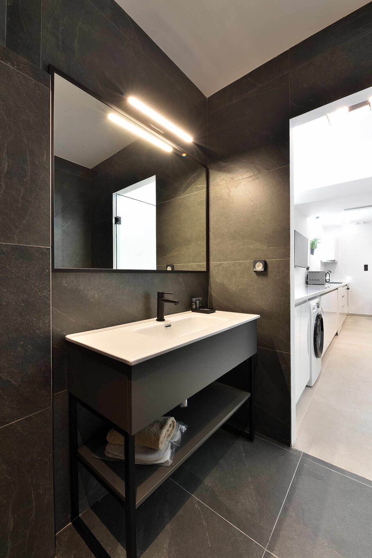 A modern bathroom with a large mirror, dark tiled walls, a white sink, and a built-in washing machine.