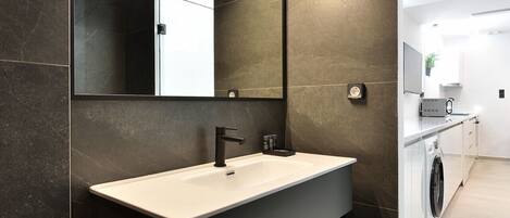 A modern bathroom with a large mirror, dark tiled walls, a white sink, and a built-in washing machine.