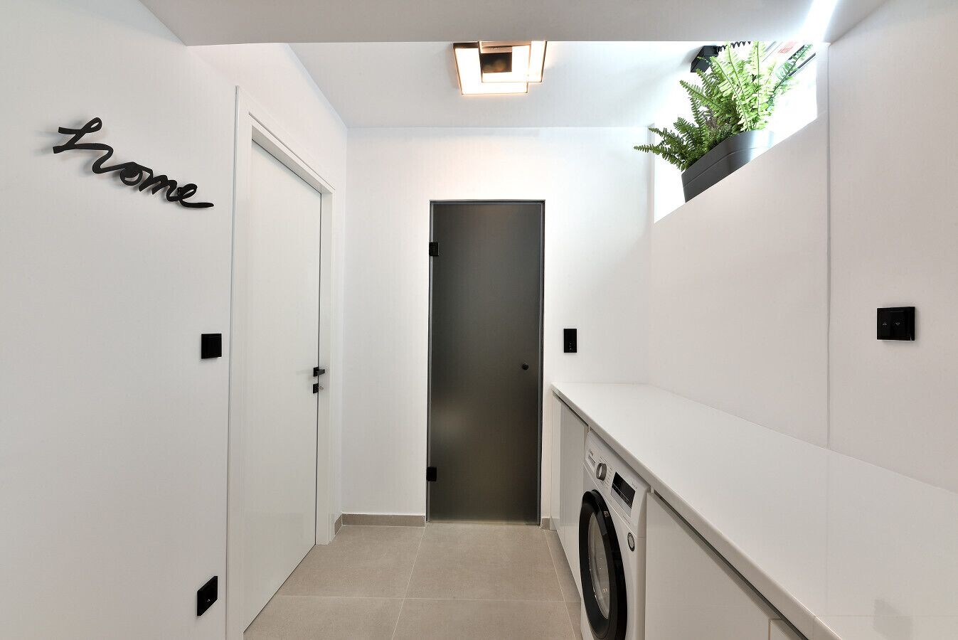 A laundry room with a washing machine, a small potted plant, and a 'home' sign on the wall.