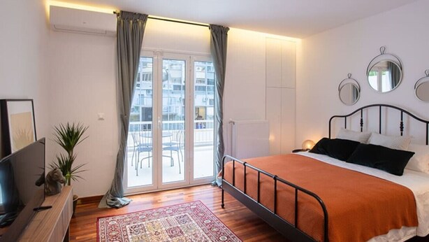 A bedroom with a bed, a TV, a plant, and a view of the outside through a glass door.