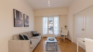 A modern living room with a sofa, a coffee table, and a chair. There are two framed artworks on the wall, a window with curtains, and a door.