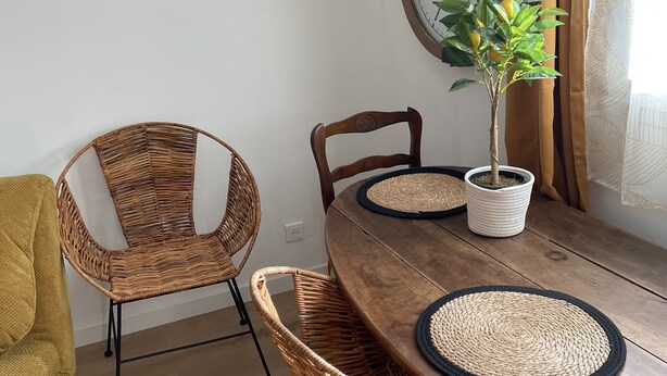 A dining area with a wooden table, wicker chairs, a potted plant, and a wall clock.