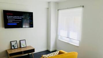 Una habitación moderna con un sofá amarillo, un televisor montado en la pared, una ventana con persiana y una pequeña mesa consola de madera con dos cuadros enmarcados.