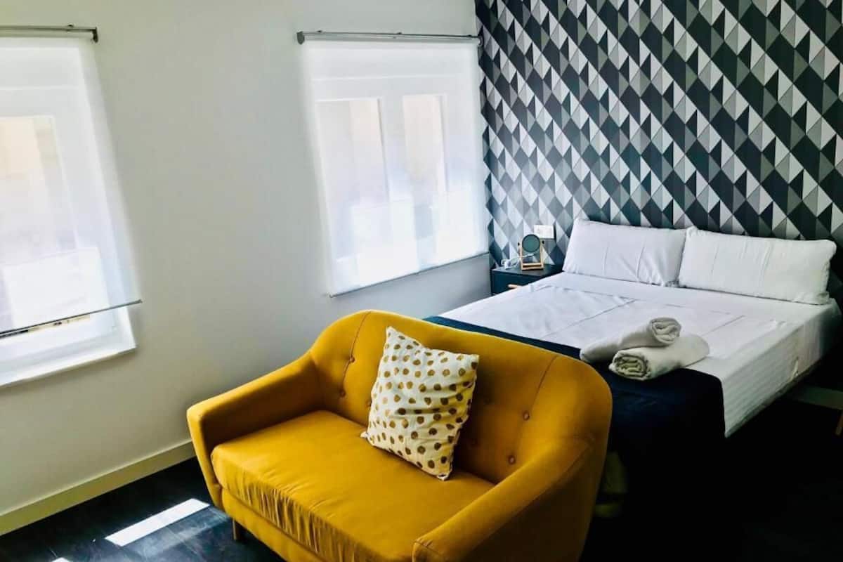 Quarto de hotel com cama, poltrona amarela com almofada de bolinhas e parede com padrão geométrico.