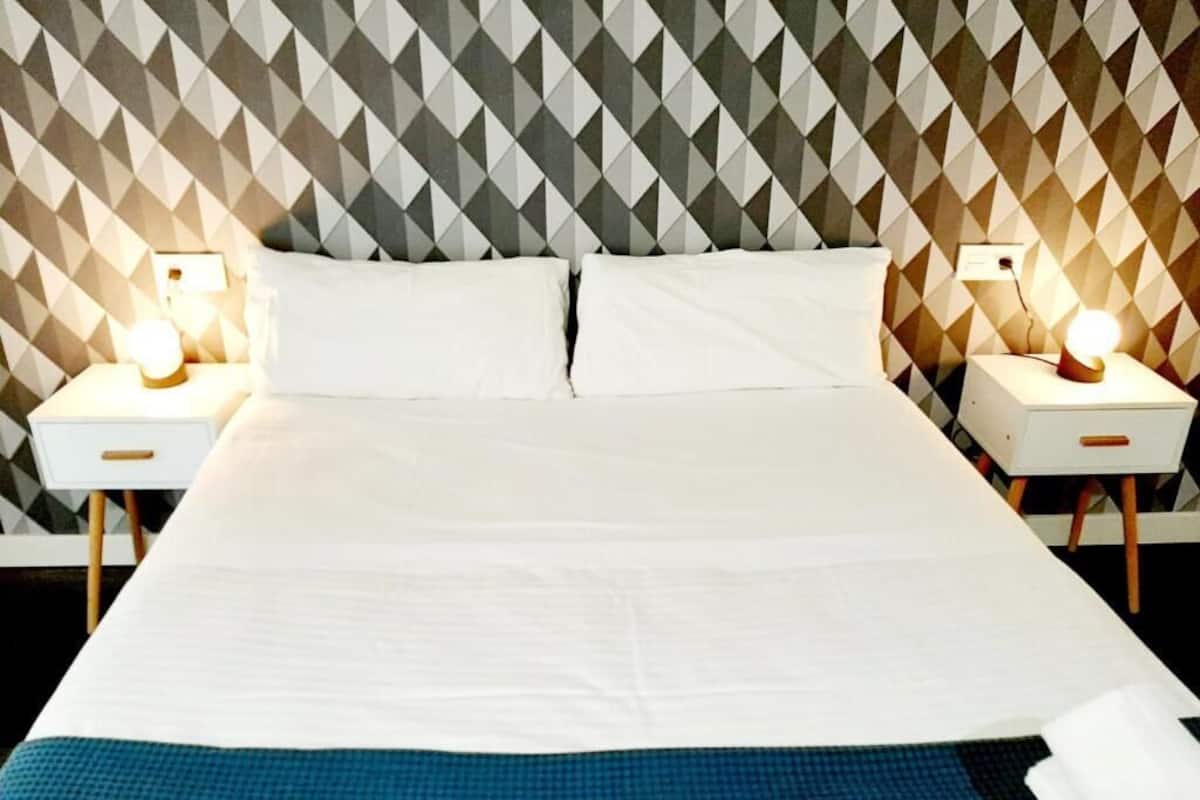 Cama bem arrumada com lençóis brancos e um toque azul no rodapé. Duas mesas de cabeceira com abajures, e a parede exibe um design geométrico.