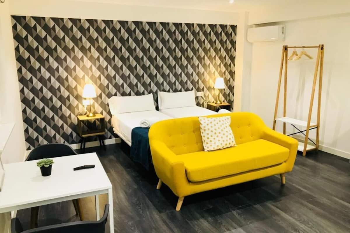 Quarto de hotel com parede estampada em padrão geométrico, sofá amarelo, cama com roupa de cama branca, duas mesas de cabeceira com abajures, escrivaninha com planta e um cabideiro de madeira.