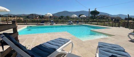 A swimming pool with lounge chairs and umbrellas, surrounded by a wooden fence and a view of houses and mountains.