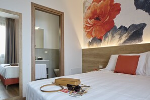 Quarto de hotel com cama, espelho, banheiro e decoração floral na parede.