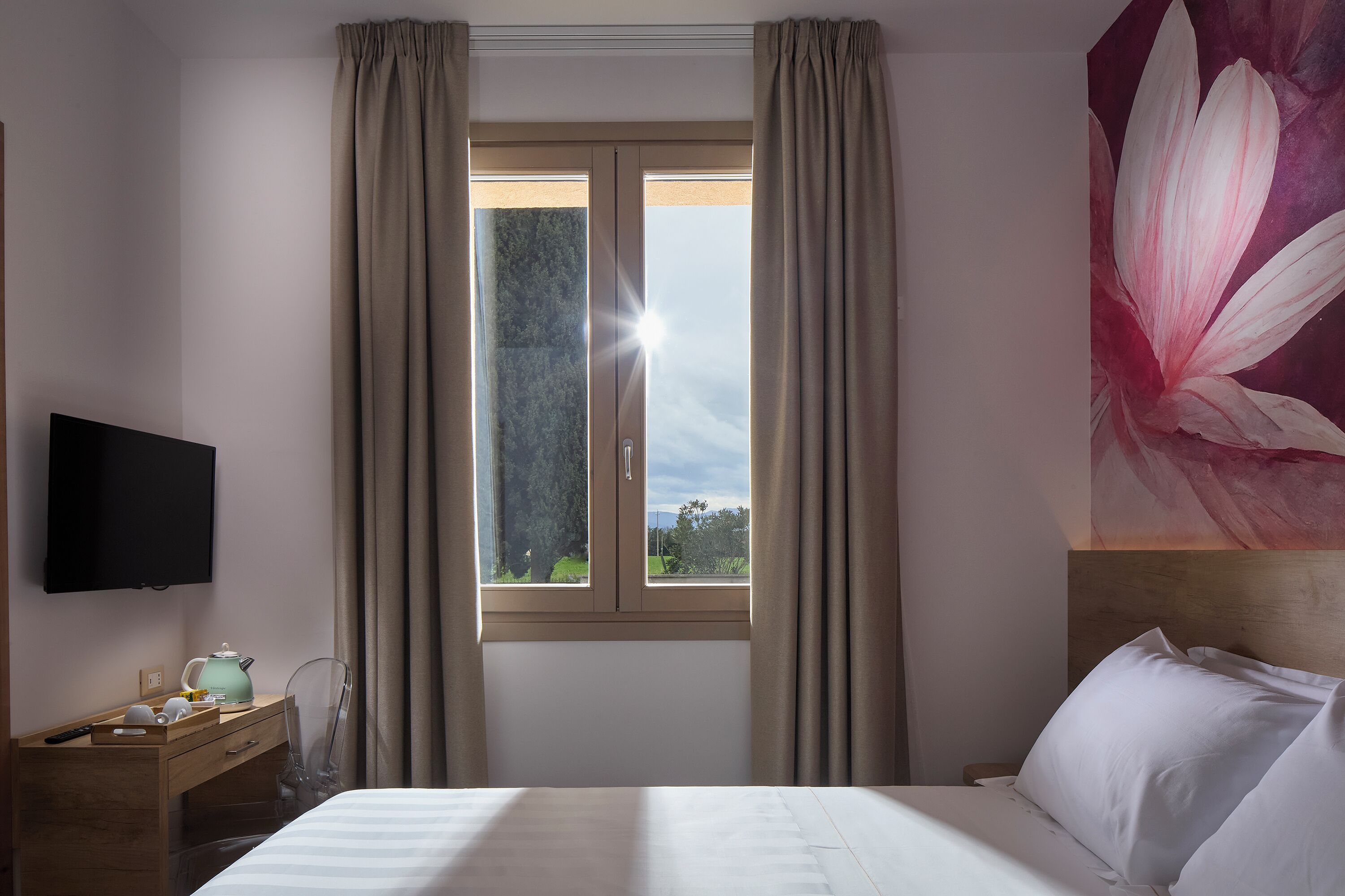 A hotel room with a bed, a desk with a kettle, a TV, and a window with curtains.