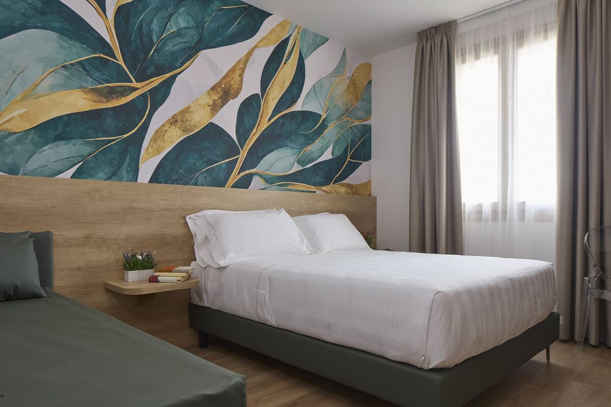 A modern hotel room with a large bed, a nightstand with a plant, a wooden headboard, and a large abstract painting on the wall.