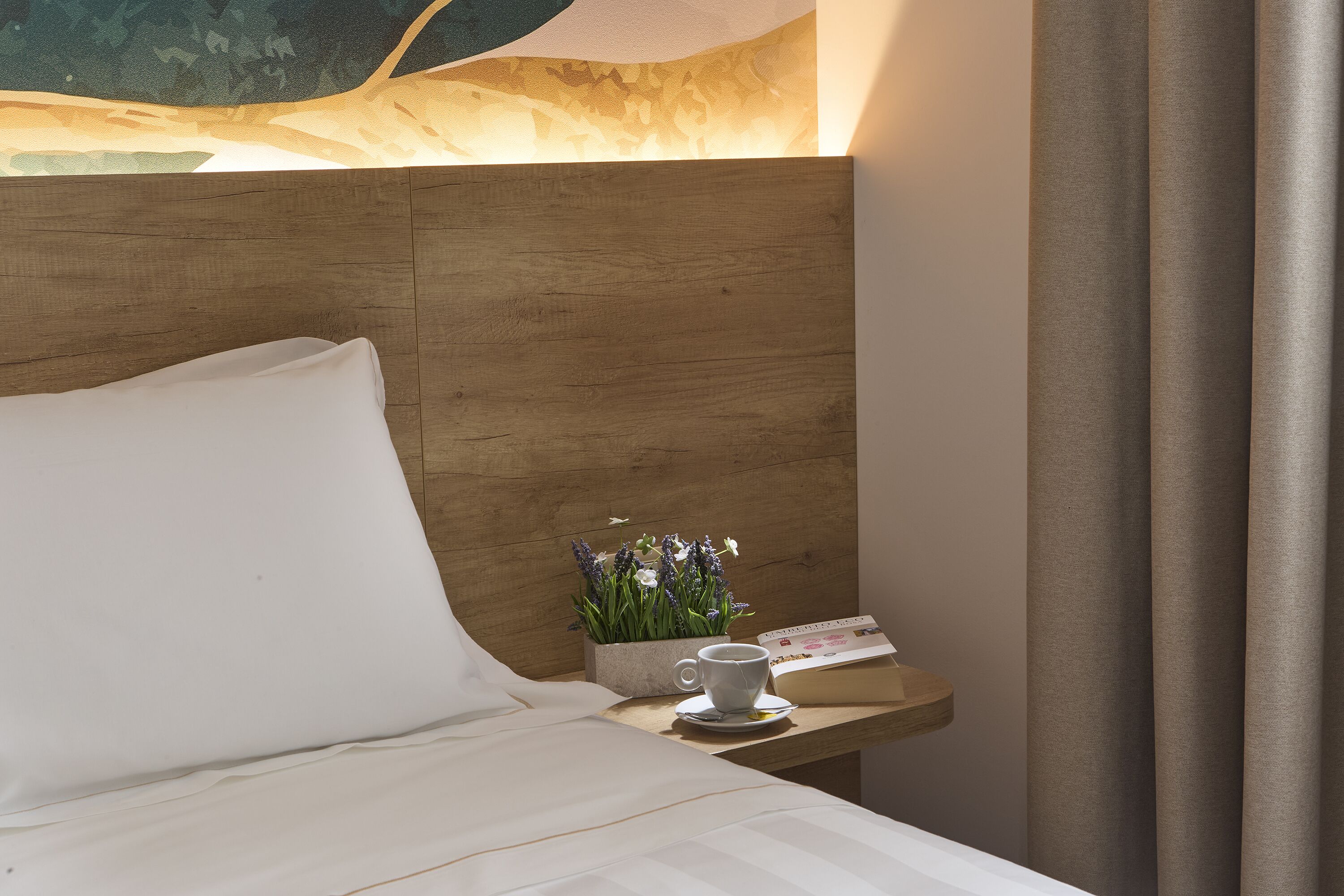 A neatly made bed with white linens, a small bedside table with a cup of coffee, a flower arrangement, and a calendar.