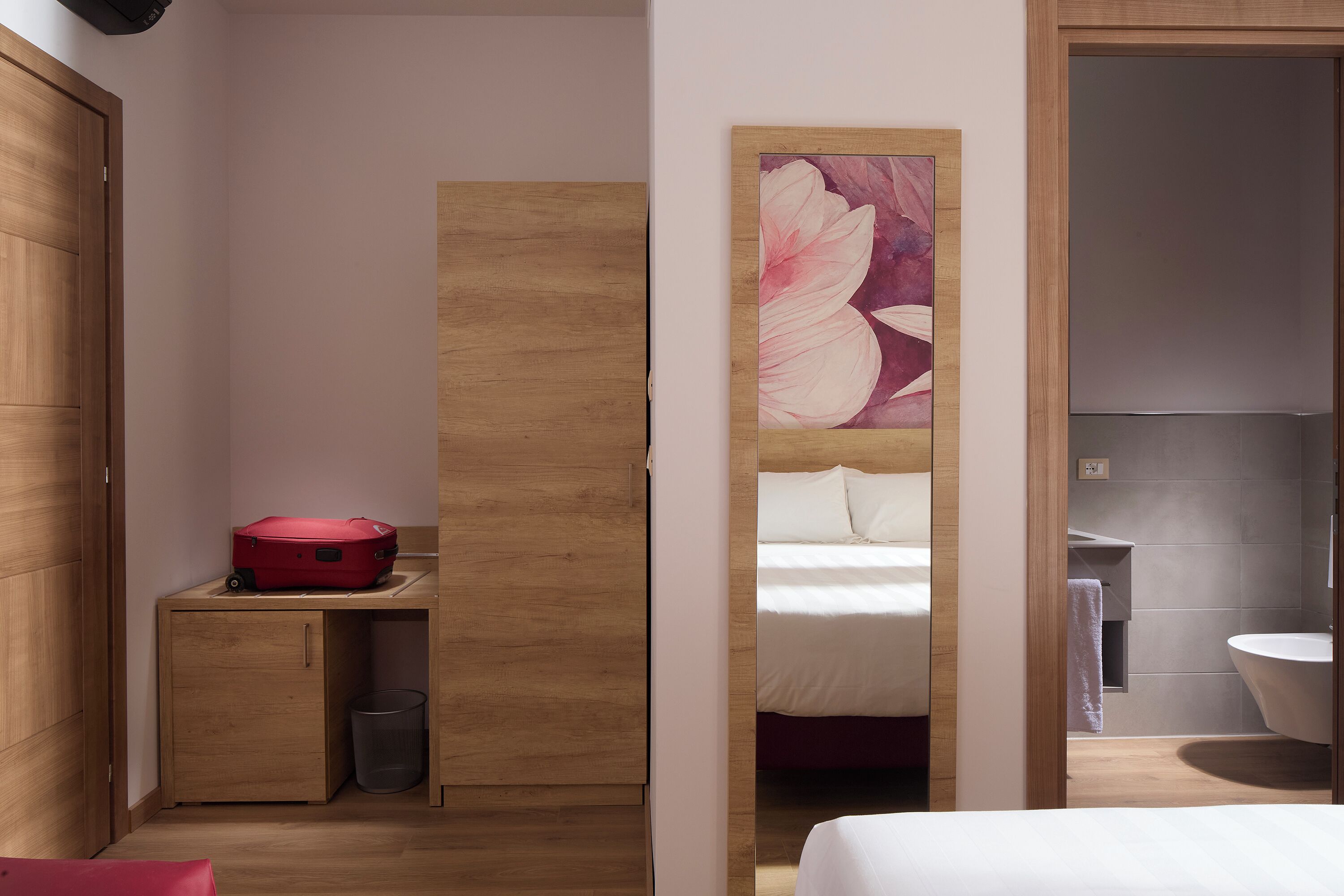 A hotel room with a wooden wardrobe, a bed with white bedding, a red suitcase on a small table, and a large floral artwork on the wall.